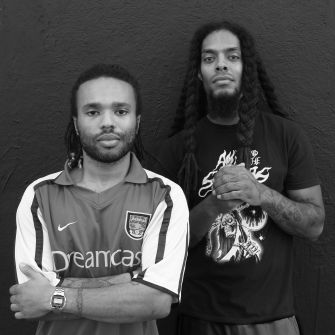 Black-and-white photo of two men standing side by side against a dark wall; the left man wears a light sports jersey with stripes, the right man wears a dark graphic T-shirt, and both look at the camera.