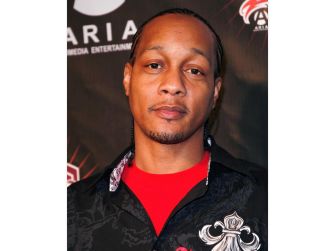 Head-and-shoulders photo of a man with braided hair wearing a red shirt and a black patterned jacket, standing in front of a dark backdrop.