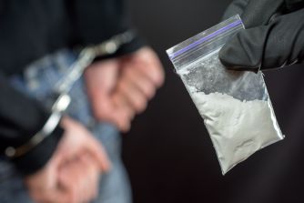 Close-up of a gloved hand holding a small zip bag containing a white powdery substance, with a blurred figure in handcuffs in the background.