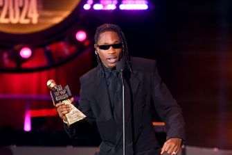 Man in a black suit and sunglasses on stage, holding a trophy award with purple lights in the background.