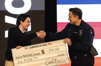 Two men on a stage shake hands while holding a large ceremonial check for one million dollars from America PAC.