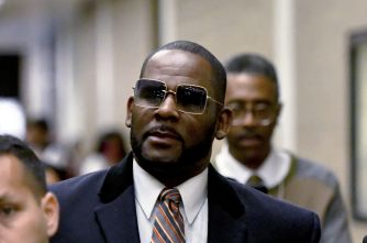 Man in a dark suit and tie wearing sunglasses, walking among people.