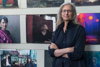 A woman with gray hair and glasses stands with arms crossed in front of a wall covered with framed photographs.