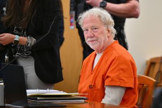 An older man with white hair wearing an orange prison jumpsuit sits at a courtroom table with papers and a laptop nearby.