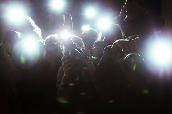 Paparazzi with cameras and bright flashbulbs in a dark setting.