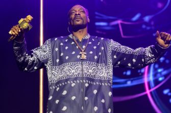 Stage performer in a blue patterned outfit with arms raised on a purple-lit stage.