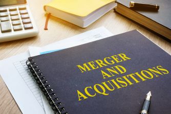 Close-up of a desk with a dark notebook titled "MERGER AND ACQUISITIONS," a calculator, books, and a pen.