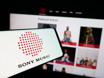 Close-up of a smartphone displaying the Sony Music logo in the foreground, with a blurred page of a music site showing artist thumbnails in the background.