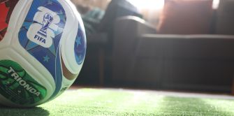 Close-up of a FIFA World Cup soccer ball on green turf, with a blurred living-room couch in the background.