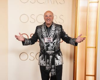Man in an elaborate black-and-silver patterned jacket poses with arms outstretched against an Oscars backdrop.