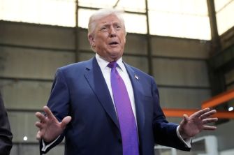 Man in a dark suit and purple tie gestures with his hands in an industrial warehouse setting.