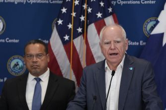 Two men in suits at a press conference, with American and state flags in the background; one man speaks at a microphone.
