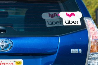 Rear view of a blue Toyota ride-hailing car with Uber and Lyft stickers on the back window and a California license plate.