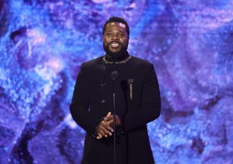 A man with dark skin wearing a black suit stands at a microphone on a stage with a blue and purple abstract backdrop.