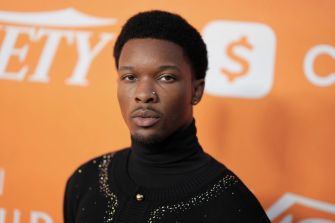 Portrait of a man in a black turtleneck with an embellished sweater, standing in front of an orange backdrop.