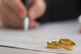 Close-up of two gold wedding bands on a document, with a hand holding a pen in the background.