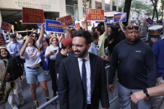 Group of protesters on a city street holding signs that read Freeze Rent, with a man in a suit walking through the crowd.