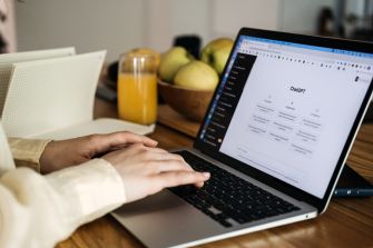 Person typing on a laptop at a wooden table with a glass of orange juice and a bowl of fruit in the background.
