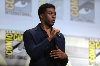 A man in a dark blazer speaks into a microphone on stage at a Comic-Con International panel, with large Comic-Con logos in the background.
