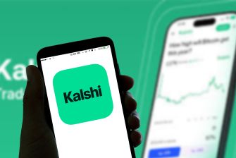Close-up of a hand holding a smartphone displaying the Kalshi app logo on a teal tile, with a second device showing a chart in the blurred background.
