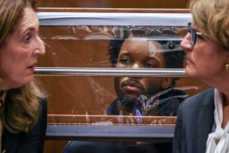 A man seated behind a glass partition in a courtroom, flanked by two women during a hearing.