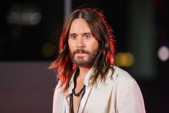 Jared Leto, with long brown hair and a beard, wearing a white shirt and necklace, stands in a dimly lit setting and looks toward the camera.