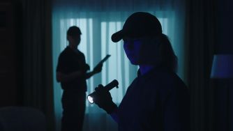 Two people in a dim, blue-lit room, a person wearing a cap in the foreground holding a flashlight while another figure stands in the background.