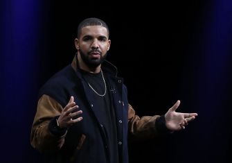 Drake, the rapper, wearing a dark jacket with tan sleeves, gestures with his hands while addressing an audience.