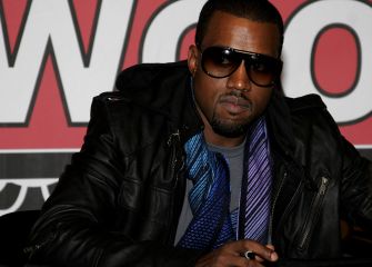 Kanye Omari West (Ye), wearing dark sunglasses and a black leather jacket with a blue scarf, sits at a table during a public appearance.