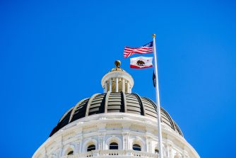 Dome of the California State Capitol with the American flag and the California state flag flying atop flagpoles against a clear blue sky.