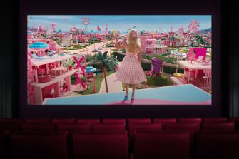 Person in a pink dress stands with their back to the camera on a theater stage, facing a large screen showing a Barbie-inspired pink cityscape.