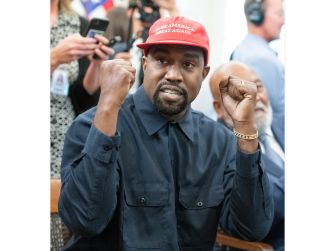 Kanye West wearing a red Make America Great Again hat and a dark blue shirt, raising his fists in the air while seated at what appears to be a public event.