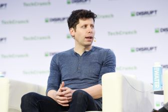 Man in a blue long-sleeve shirt sits on a stage chair with a headset microphone, speaking at a tech conference with sponsor logos in the background.