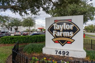Taylor Farms sign with the company logo reading “Taylor Farms Finest Quality Salads” in a landscaped area, with a row of parked cars and a building in the background.