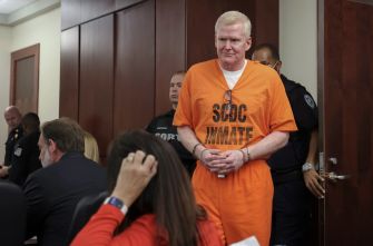 Alex Murdaugh, wearing an orange jail jumpsuit labeled “SCDC INMATE,” stands in a courtroom as observers and security personnel look on.