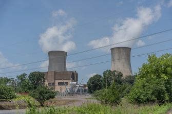 A nuclear power plant with two large cooling towers releasing steam, with trees in the foreground and power lines overhead.