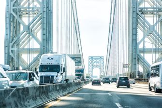 Traffic on the George Washington Bridge's upper deck, including several tractor-trailers and cars, with the bridge's steel framework visible overhead.