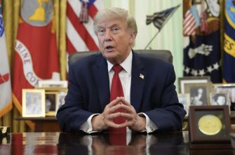Donald Trump sits at a polished desk in a formal office, hands clasped on the desk, with U.S. flags and framed photos in the background.