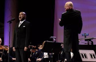 Jubilant Sykes performs onstage in a tuxedo, singing into a microphone with an orchestra behind him.