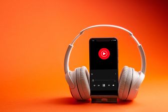 Smartphone with a red play button on its screen is flanked by white over-ear headphones on an orange background, symbolizing music streaming and licensing related to the Downtown Music–Blast Off dispute.
