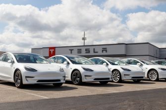 Row of white Tesla electric cars parked in front of a Tesla dealership with a large TESLA sign on the building.