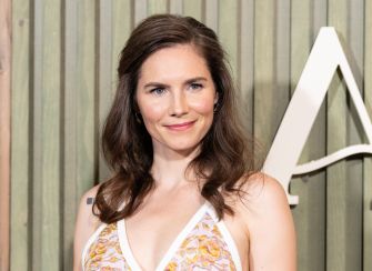 Amanda Knox poses on a red carpet, smiling, wearing a white sleeveless top with a floral pattern.