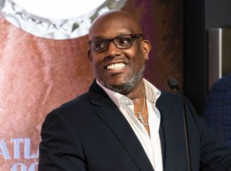Wayne Barrow, Notorious B.I.G.’s longtime manager, wearing a dark suit and white shirt with a cross necklace, smiles at a public event.