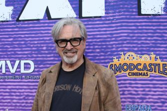 Robert Carradine poses for a photo at an event, standing in front of a purple backdrop with logos while wearing a brown suede jacket, a dark T-shirt, and glasses.