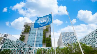 A United Nations flag flutters atop a flagpole in front of a modern glass office complex under a bright blue sky.