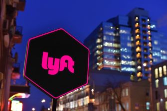 Lyft logo sign glowing pink on a city street at night with tall buildings in the background.