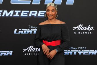 Tiffany Haddish poses for photos on a carpet in an off-the-shoulder black outfit with a bright red belt, in front of a backdrop with promotional logos.