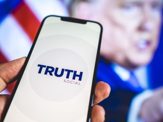 Smartphone screen shows the Truth Social logo, held up with a blurred figure giving a speech in the background.