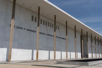 Exterior view of the John F. Kennedy Center for the Performing Arts in Washington, D.C., featuring a long white façade with tall vertical columns.