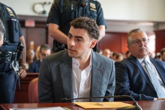 Luigi Mangione sits at a courtroom table in a gray suit and white shirt during a federal plea hearing, with security officers visible in the background.